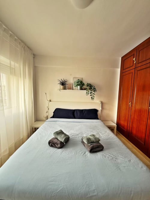 Luminoso dormitorio de apartamento de playa en Fuengirola con cama blanca, almohadas oscuras, armario de madera y plantas en macetas