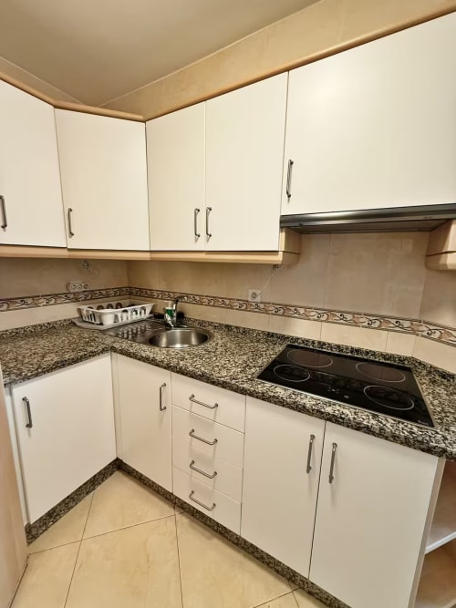 Cocina moderna con muebles blancos, encimeras de granito, placa eléctrica y fregadero de acero inoxidable en apartamento de Fuengirola