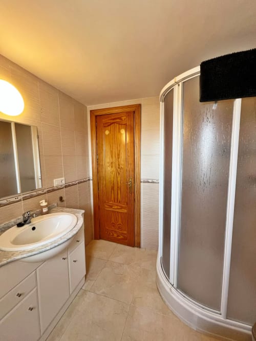 Baño moderno con tocador blanco, lavabo redondo, espejo, puerta de madera y ducha curva en apartamento de Fuengirola