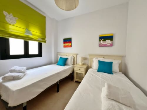 Dormitorio doble en apartamento de playa en Fuengirola con almohadas turquesas, muebles crema y persianas verdes lima