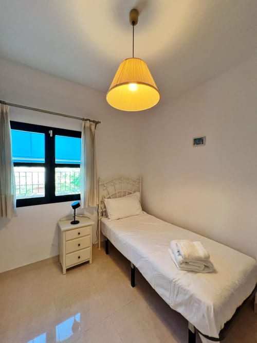 Dormitorio luminoso en apartamento de Fuengirola con cama individual, mesita de noche de madera, lámpara colgante y vistas a la ciudad