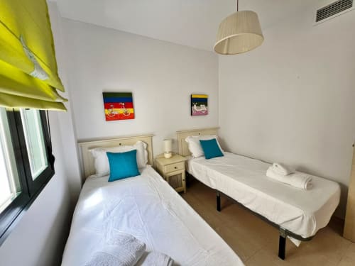 Dormitorio doble luminoso en apartamento de playa en Fuengirola con ropa de cama blanca, almohadas turquesas, luz natural y pared colorida