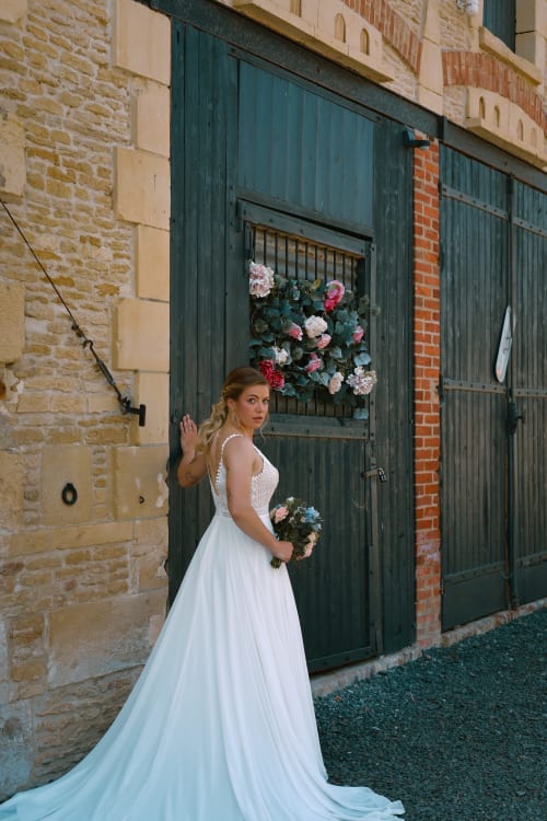 Mariée en robe blanche posant à l'entrée en pierre du Domaine Du Grand Fort, décorée de fleurs, en Normandie
