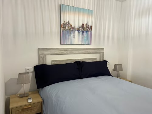 Dormitorio de lujo moderno en apartamento de playa en Fuengirola con obra de arte de vista urbana, cabecero gris, almohadas negras y lámparas laterales