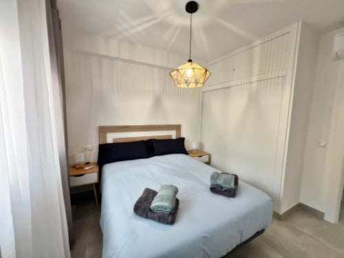 Dormitorio moderno en Grupo Inmo House Fuengirola con cama queen, almohadas negras, toallas dobladas, lámpara colgante y diseño minimalista
