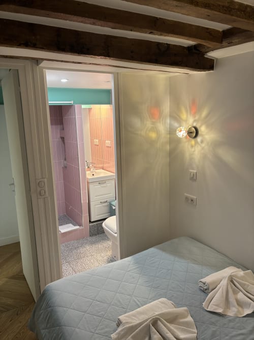 Modern bedroom in Paris apartment with ensuite bathroom, exposed beams, and decorative wall sconce lighting