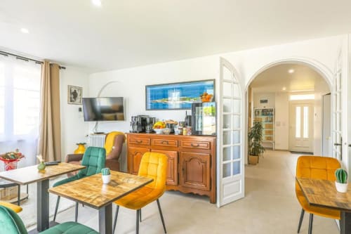 Modern dining and kitchen area at La Villa Cassinne B&B with yellow chairs, wooden table, orange dresser, and view through