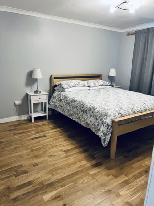 Spacious bedroom with wooden bed, white nightstand, lamps, and hardwood floors at Whiteley's Cottages cottage in Dumbarton