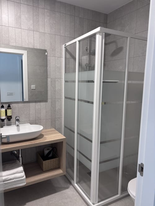 Baño moderno en La Llar de Carboneras con azulejos grises, ducha blanca, lavabo de cerámica y mueble de madera