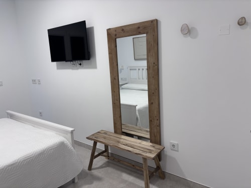 Modern double bedroom at La Llar B&B in Carboneras with wooden-framed mirror, wooden bench, flat-screen TV, and white bedding