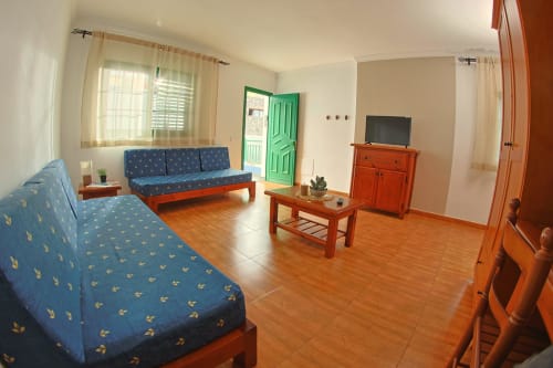 Luminosa sala de estar en Apartamentos La Palmera con sofás azules, muebles de madera, TV, puerta verde y vistas al mar desde la terraza