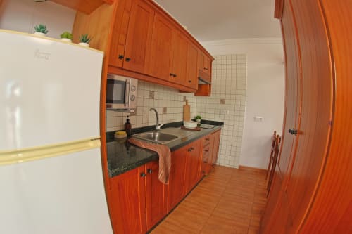 Cocina compacta con gabinetes de madera naranja, fregadero de acero inoxidable y backsplash de azulejos blancos en Apartamentos La Palmera