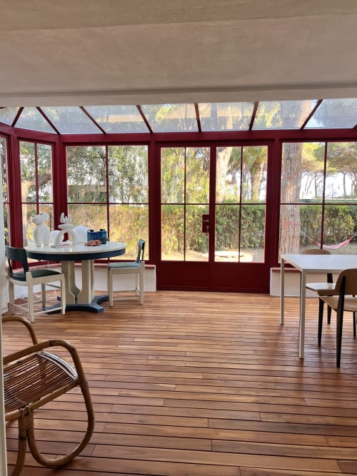 Sunlit villa dining area with wooden floors, red-framed windows, sea views, round table and chairs at Casa Pineta