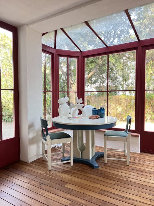 Modern dining area in Casa Rossa villa with round white table, burgundy-framed windows overlooking Marina di Campo gardens