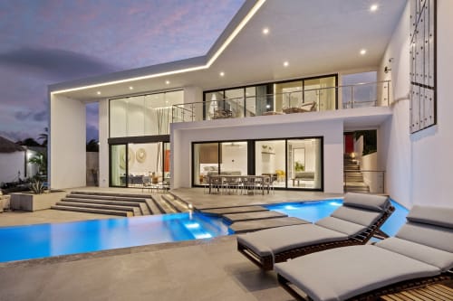 Luxury villa at Isla Mujeres Realty with illuminated pool, modern architecture, ocean views, and lounge seating at dusk
