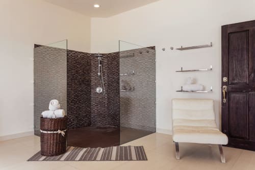 Modern bathroom with walk-in shower, glass enclosure, dark tile, floating shelves, wicker basket, and cream lounge chair at