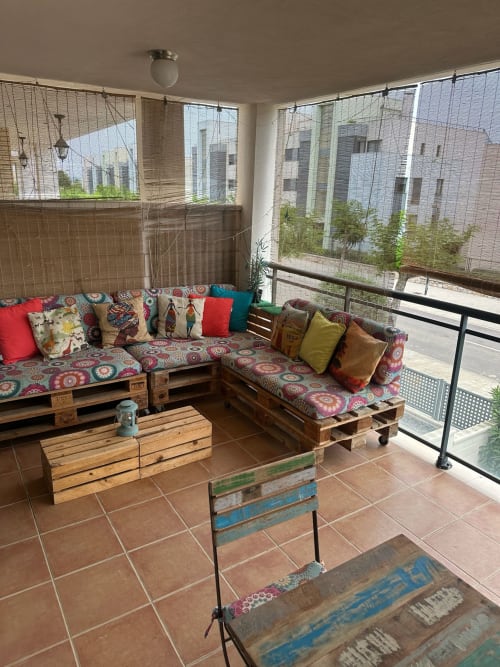 Terraza cerrada acogedora con muebles de palet, cojines coloridos, vistas a las montañas y edificios de ladrillo visibles a través de malla