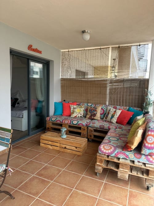 Terraza acogedora de apartamento en Sant Jordi con muebles de palés, cojines coloridos y vistas a las montañas a través de mallas