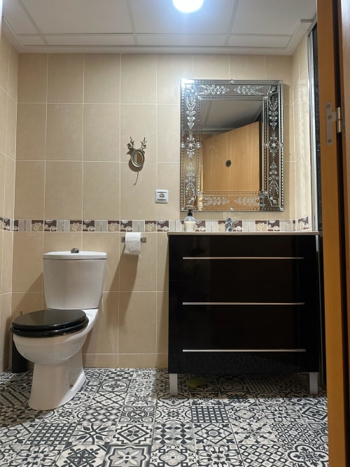 Baño moderno del apartamento Sant Jordi con azulejos beige, tocador negro, espejo decorativo, inodoro y piso con patrón