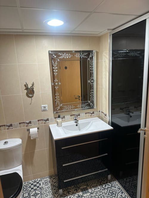 Baño moderno del apartamento Sant Jordi con lavabo negro, espejo ornamentado, azulejos beige, inodoro, área de ducha y decoración