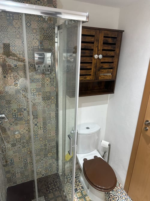 Baño moderno con ducha de azulejos decorativos, inodoro blanco, mueble de madera y espejo en apartamento Sant Jordi