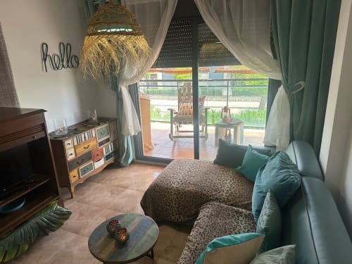Acogedora sala de estar del apartamento Sant Jordi con terraza, lámpara de mimbre, cortinas verdes, ropa de cama leopardo y vista a la montaña