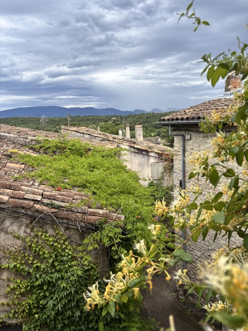 Bâtisse en pierre rustique aux toits en terre cuite surplombant la campagne française verdoyante et les montagnes lointaines sous un ciel nuageux
