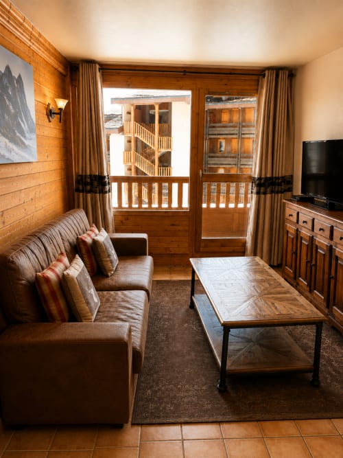 Warm wood-paneled living room at Le Chalet des Cimes with leather sofa, coffee table, TV, and balcony overlooking Belle
