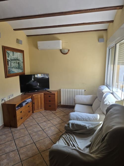 Sala de estar de la casita Oliva con sofá gris, mueble de madera para TV, paredes amarillas y techo con vigas