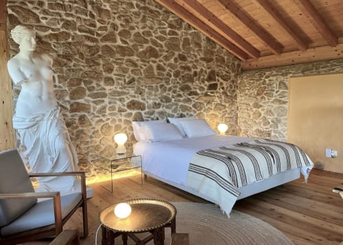 Quarto na Quinta da Mata de Baixo com paredes de pedra, vigas de madeira, cama de casal, busto clássico e área de estar.