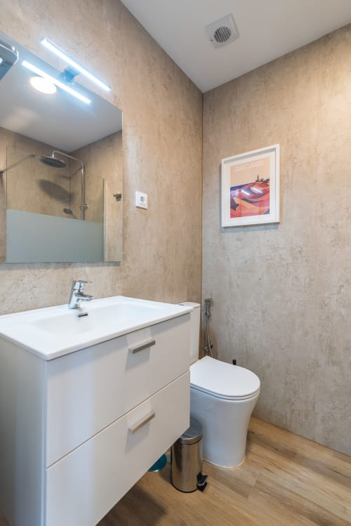 Modern bathroom at Home Stay Gran Canaria with white vanity, toilet, travertine tile walls, and Gran Canaria poster