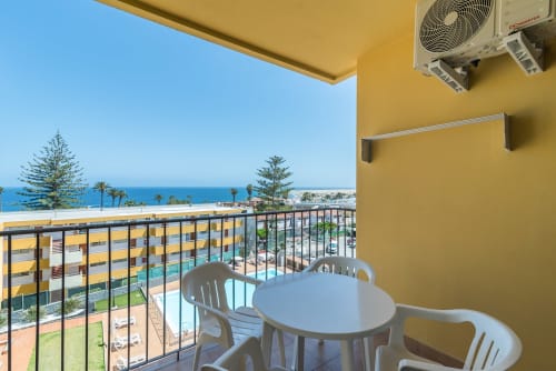 Beachfront balcony with sea views at Home Stay Gran Canaria, showing white furniture, pool, and palm trees overlooking the