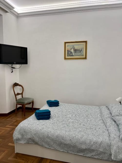 Camera moderna a Domus Giovanna Roma con letto matrimoniale, asciugamani blu, sedia in legno, TV schermo piatto e quadri su muri bianchi