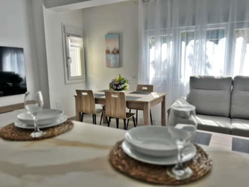 Moderna zona de comedor y estar diáfana en Fuengirola con mesa servida, sillas de madera, sofá gris e iluminación natural