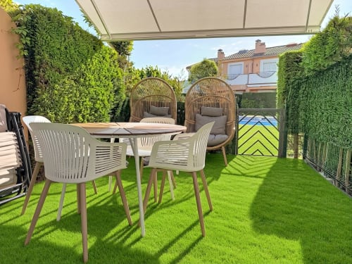 Patio soleado de la casa de alquiler Estartit con mesa de comedor, sillas de mimbre, césped artificial, setos verdes y toldo retráctil
