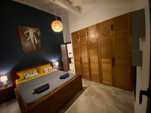 Chambre moderne avec lit en bois, coussins jaunes, murs bleu marine, lampe suspendue et grand dressing en bois aux Hébergements