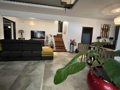 Séjour moderne ouvert au gîte Les Hébergements Passion avec canapé noir, téléviseur, escalier en bois, coin repas et verdure