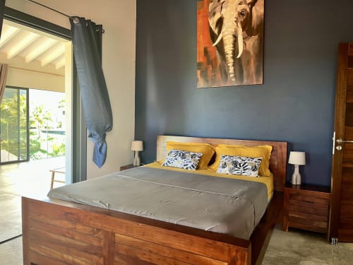 Chambre de la maison Les Trois-Ilets avec lit en bois, murs bleus, coussins jaunes, œuvre d'art éléphant et accès à la vue mer