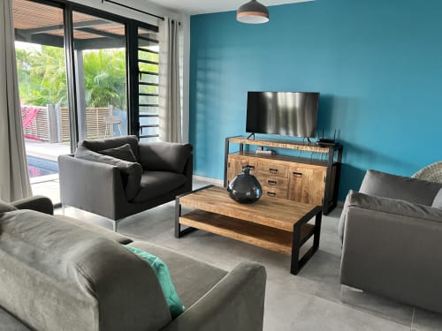 Salon moderne aux murs turquoise, canapés gris, table basse en bois, téléviseur sur meuble bois, vue sur la mer par grandes fenêtres