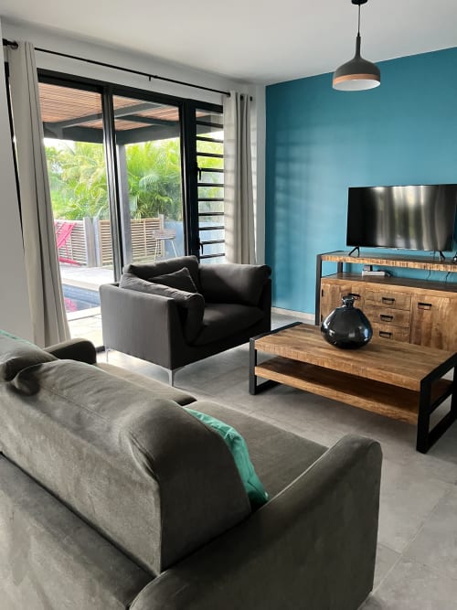 Salon moderne avec mur d'accent turquoise, canapé gris, fauteuil noir, TV écran plat, table basse en bois et portes avec vue sur mer
