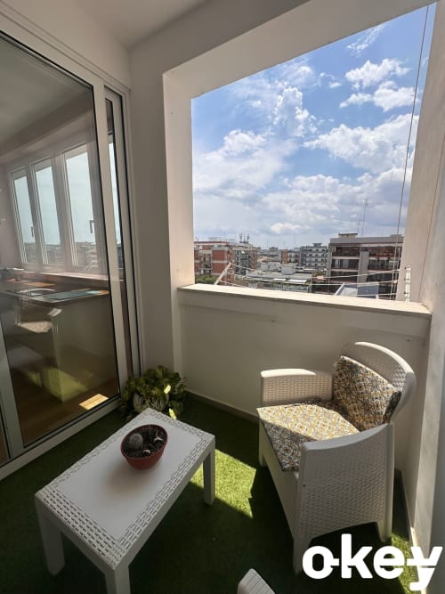 Balcone soleggiato con area salotto con vista panoramica su Bari, pianta in vaso su tavolo bianco, erba sintetica verde