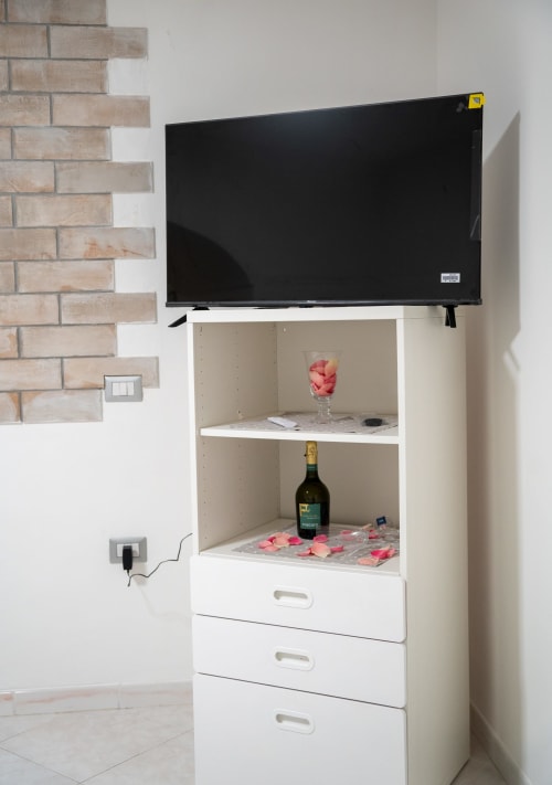 White TV stand with shelves displaying wine bottle and flowers, flat-screen TV mounted above in Sorrento apartment