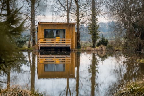 Cabane en bois aux Rives du bel air reflétée dans une eau calme, entourée d'arbres nus et de végétation humide
