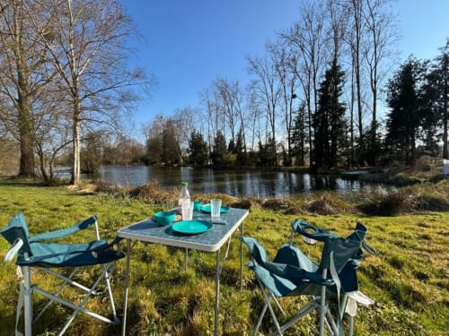 Aménagement pique-nique au bord de la rivière aux Rives du bel air avec chaises de camping, vaisselle turquoise et arbres dénudés se reflétant dans l'eau calme