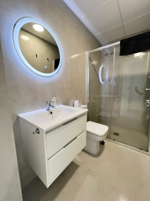 Baño de lujo moderno en apartamento de Fuengirola con espejo redondo iluminado LED, lavamanos blanco, inodoro y ducha de cristal