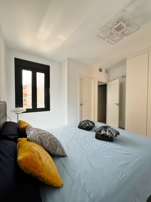 Dormitorio moderno en apartamento de lujo en Fuengirola con ventana de marco negro, cama azul y cojines amarillos