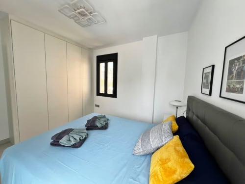 Dormitorio de lujo moderno en apartamento de Fuengirola con ropa de cama azul, cabecero gris, almohadas amarillas y armario blanco.