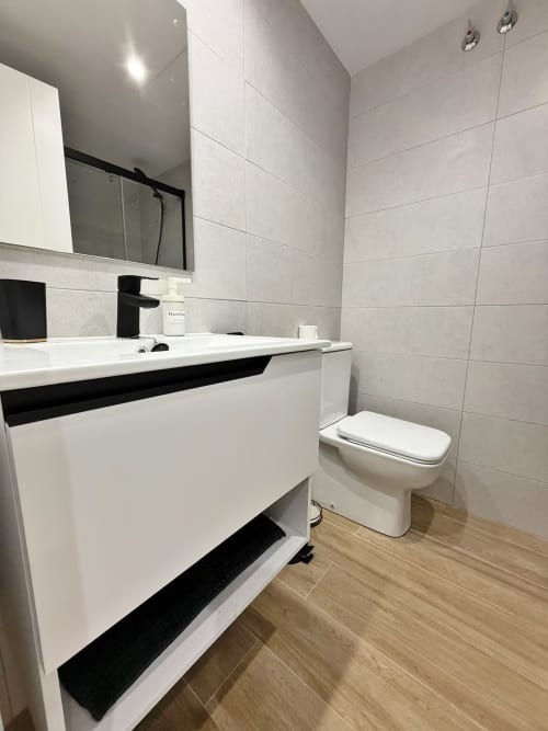 Baño moderno en apartamento frente al mar en Fuengirola con lavamanos blanco, grifo negro, inodoro y suelo efecto madera
