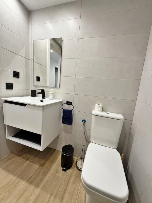 Cuarto de baño moderno en apartamento frente al mar, Fuengirola. Lavabo blanco, inodoro, espejo iluminado, azulejos grises, efecto madera