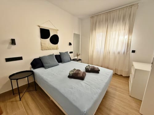 Dormitorio moderno en apartamento de playa en Fuengirola con cama blanca, acentos negros, suelos de madera y grandes ventanales con cortinas